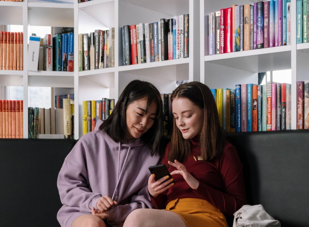 Dve mladé dievčatá hľadiace do mobilu sediace na čiernej pohovke pred bielou knižnicou plnou kníh